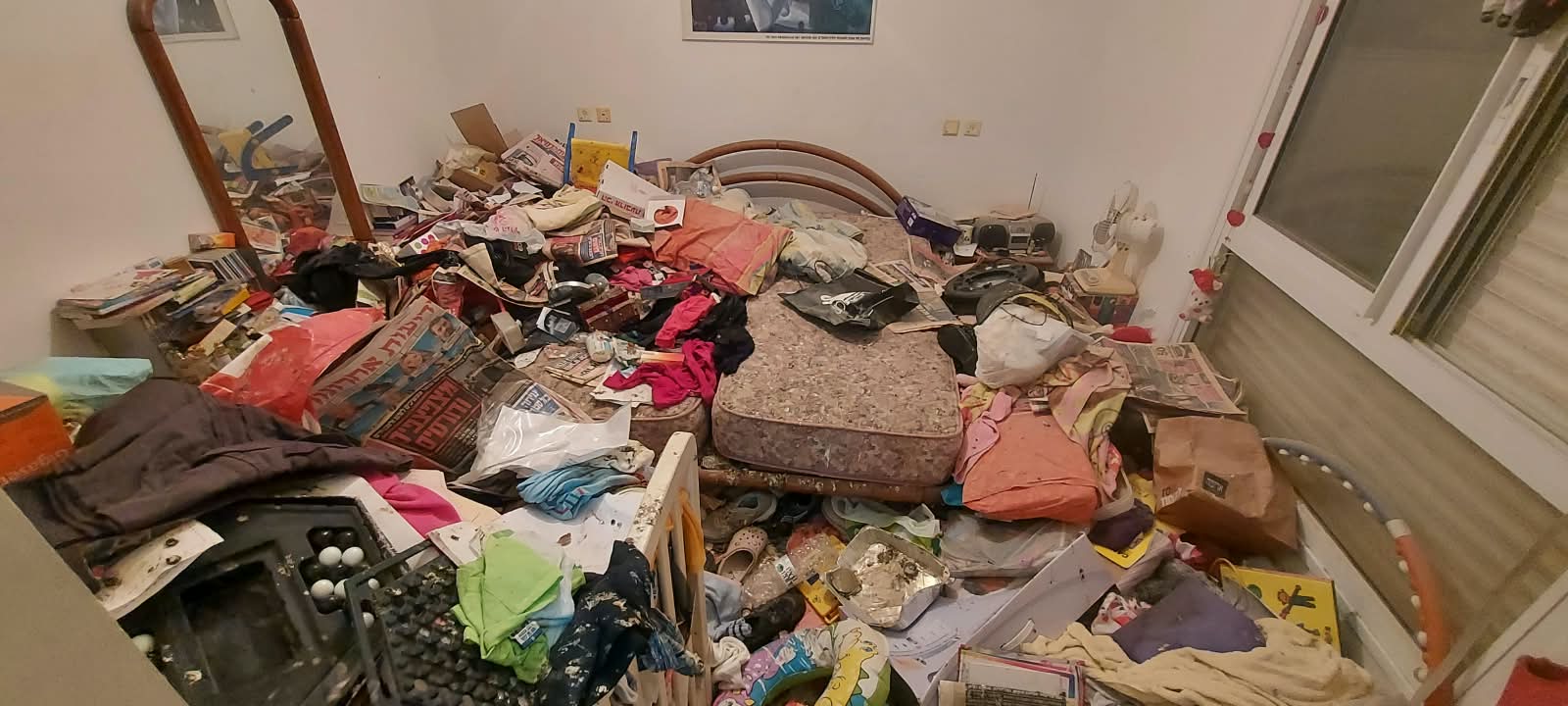 חדר שינה עמוס בעיתונים, בגדים וחפצים לפני פינוי